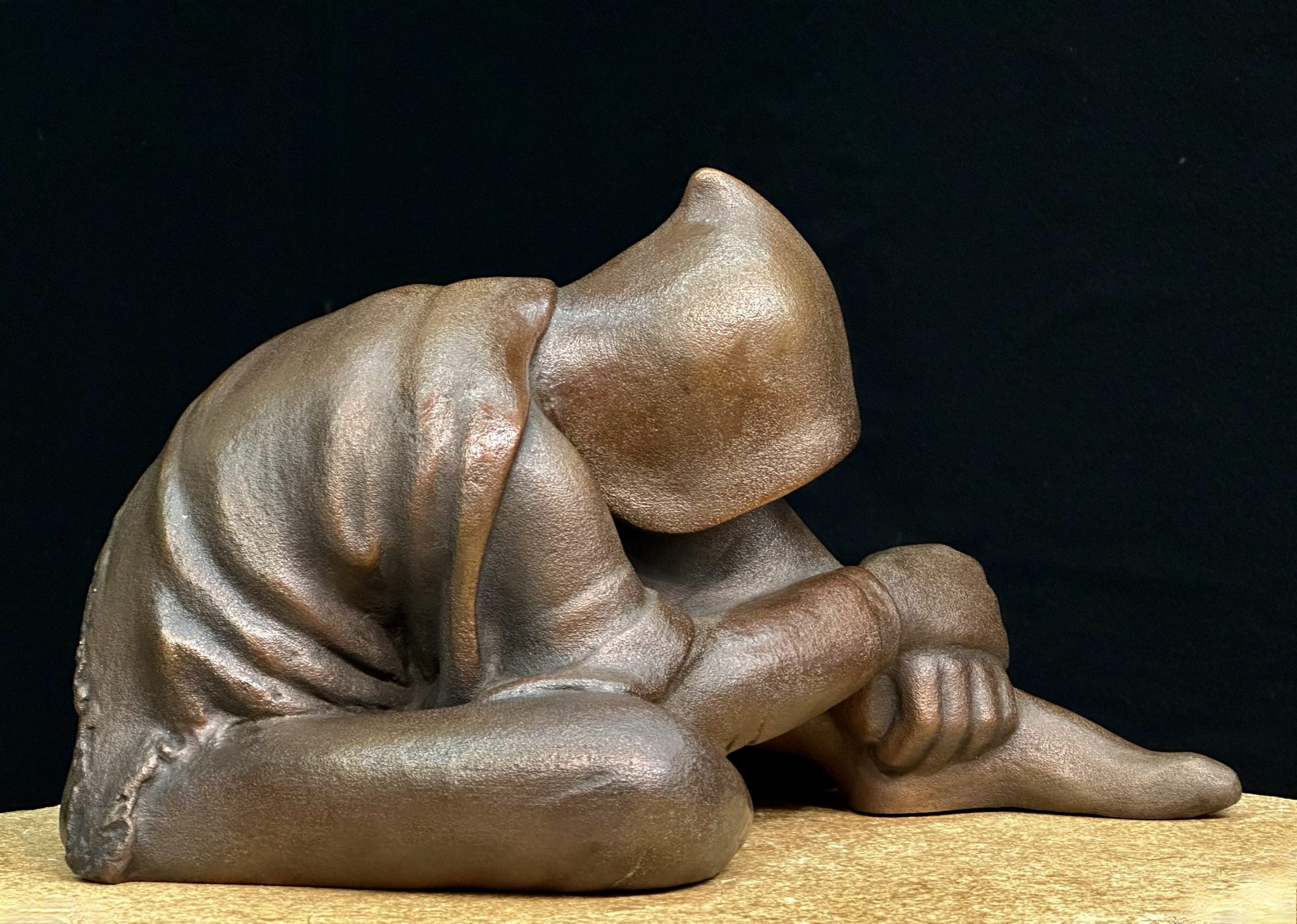 A bronze sculpture of an anonymous figure huddling forward, the right leg bent and externally rotated, the left leg bent with knee upright with both arms wrapped about it. The figure is draped with what appears to be wearing a hooded sweatshirt and is draped with a ragged blanket.
